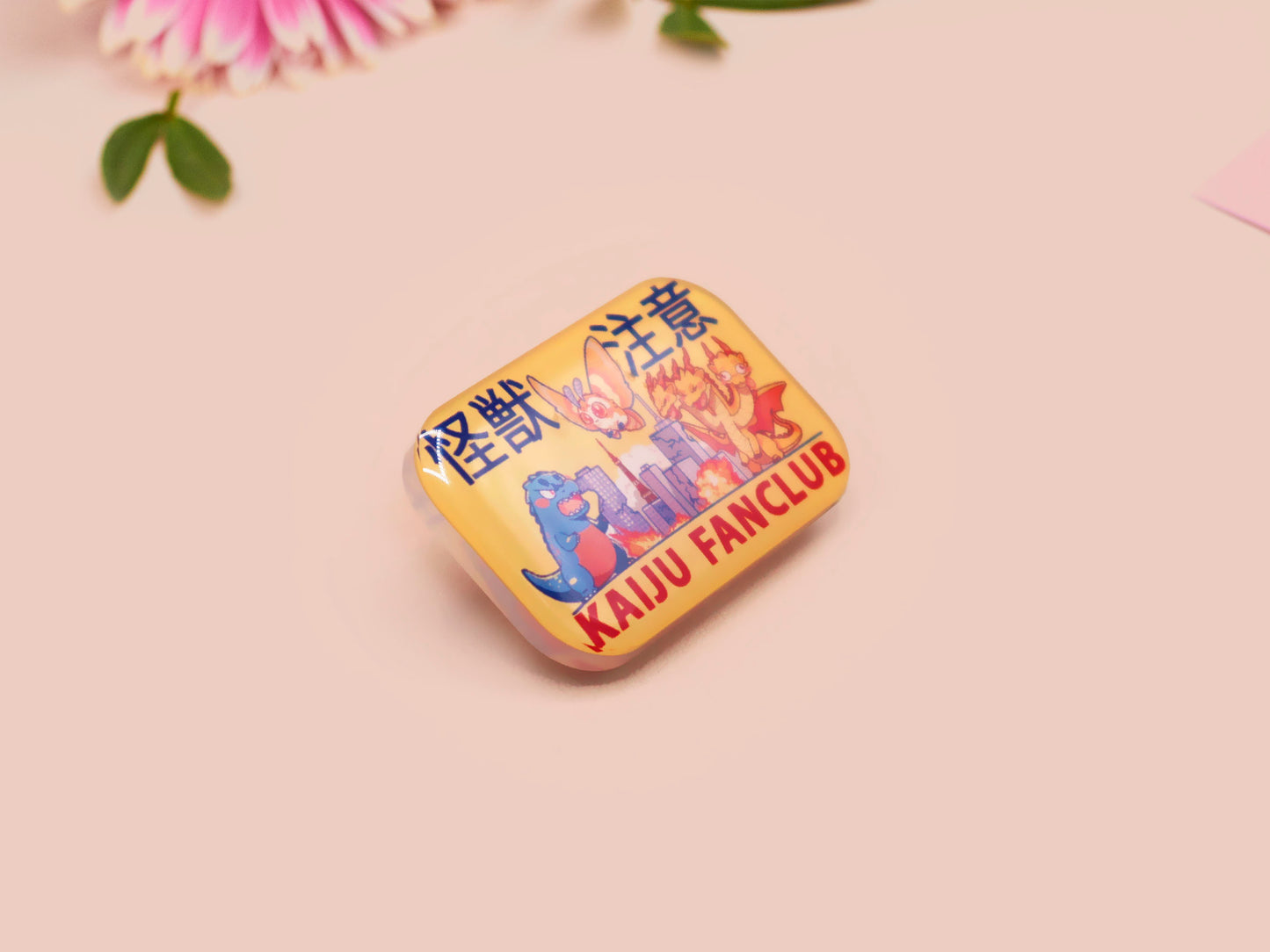 A clear acrylic pin badge with a pink heart-shaped pin back clasp. The design of the pin badge is of a cute chibi blue godzilla like kaiju mothra and King Ghidorah destroying a city with the quote kaiju fanclub and kaiju warning in japanese kanji