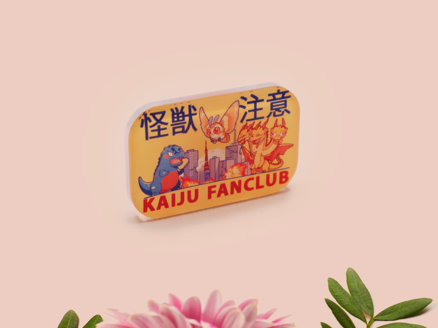A clear acrylic pin badge with a pink heart-shaped pin back clasp. The design of the pin badge is of a cute chibi blue godzilla like kaiju mothra and King Ghidorah destroying a city with the quote kaiju fanclub and kaiju warning in japanese kanji