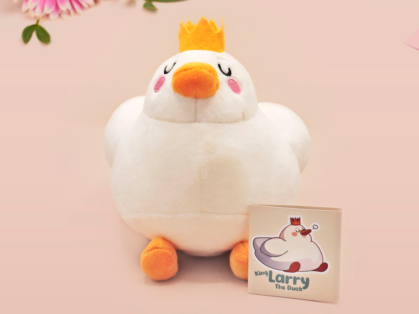 An adorable white duck bird plushie toy with cute embroidered eyes and facial expression, wearing a yellow crown hat, with a hang tag describing the duck as King Larry.