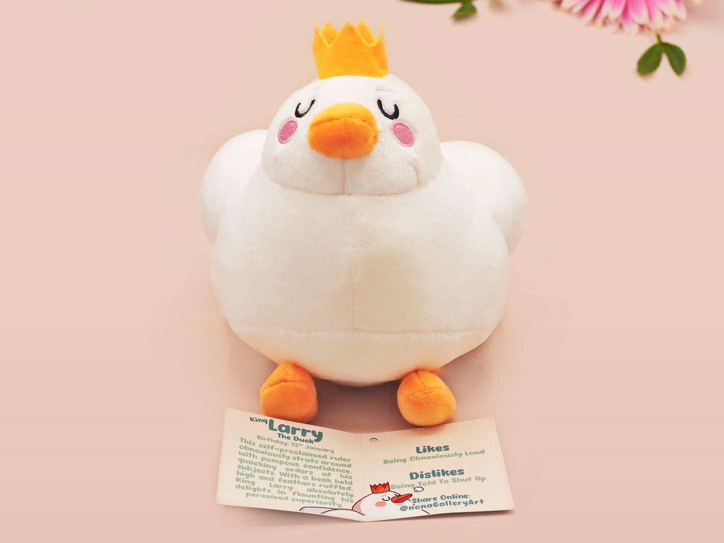 An adorable white duck bird plushie toy with cute embroidered eyes and facial expression, wearing a yellow crown hat, with a hang tag describing the duck as King Larry.
