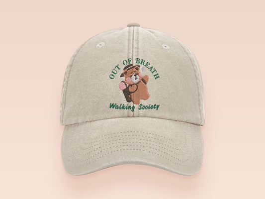 A cream baseball cap hat with an embroidered cute brown bear hiking with the quote out of breath walking society.