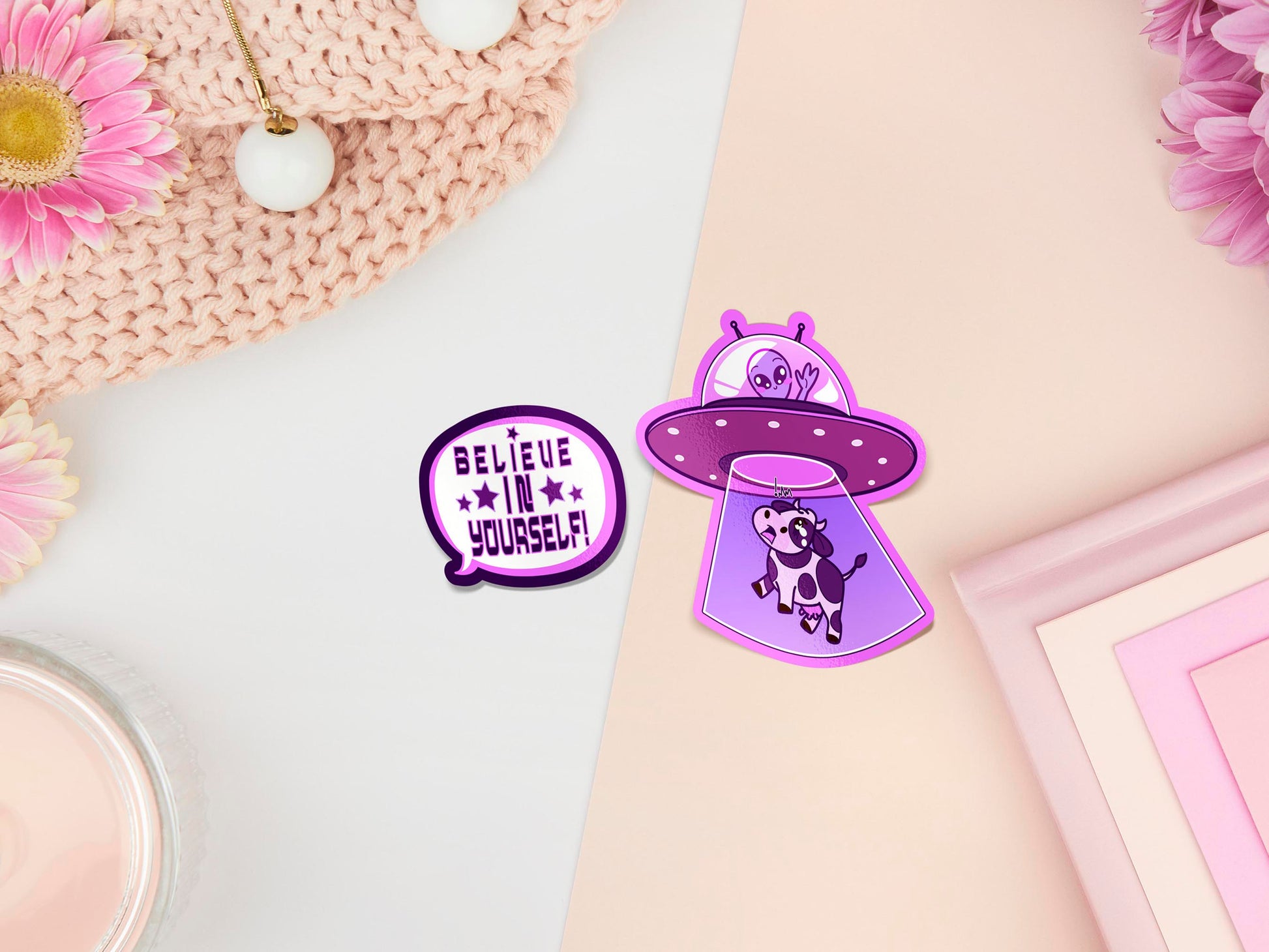 Two stickers of digitally illustrated cartoons one is a speech bubble with the words Believe in yourself. The other sticker is a happy alien in a UFO abducting a shocked cow