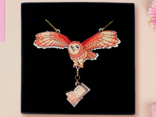 Mixed material handmade necklace of chibi cartoon flying barn owl with pearlescent wings carrying a removeable spell book charm, with a gold chain and black gift box.