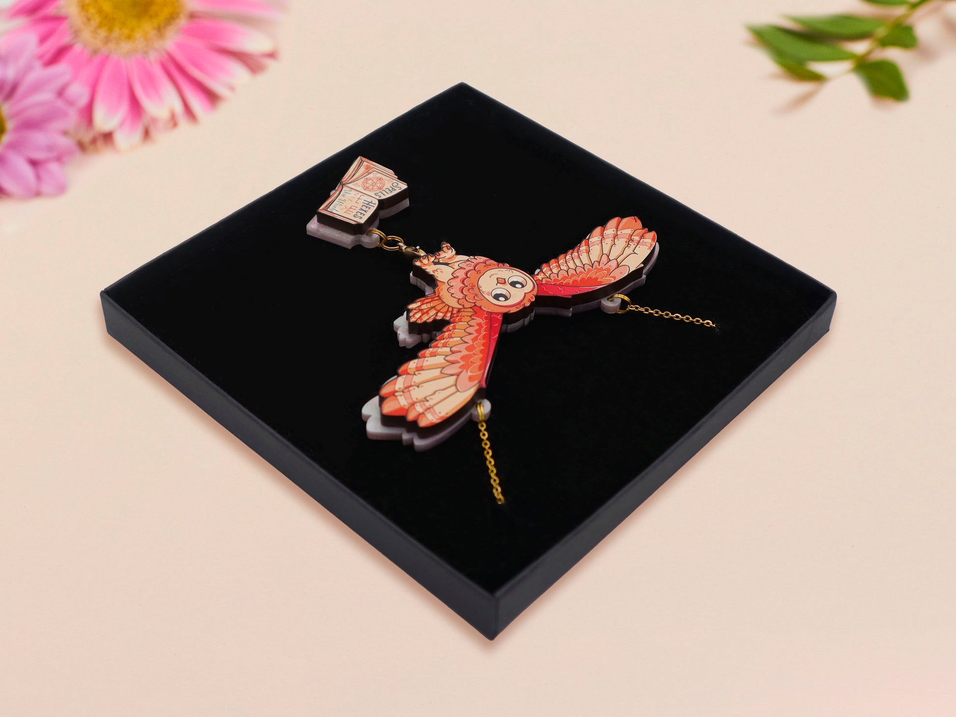 Mixed material handmade necklace of chibi cartoon flying barn owl with pearlescent wings carrying a removeable spell book charm, with a gold chain and black gift box.