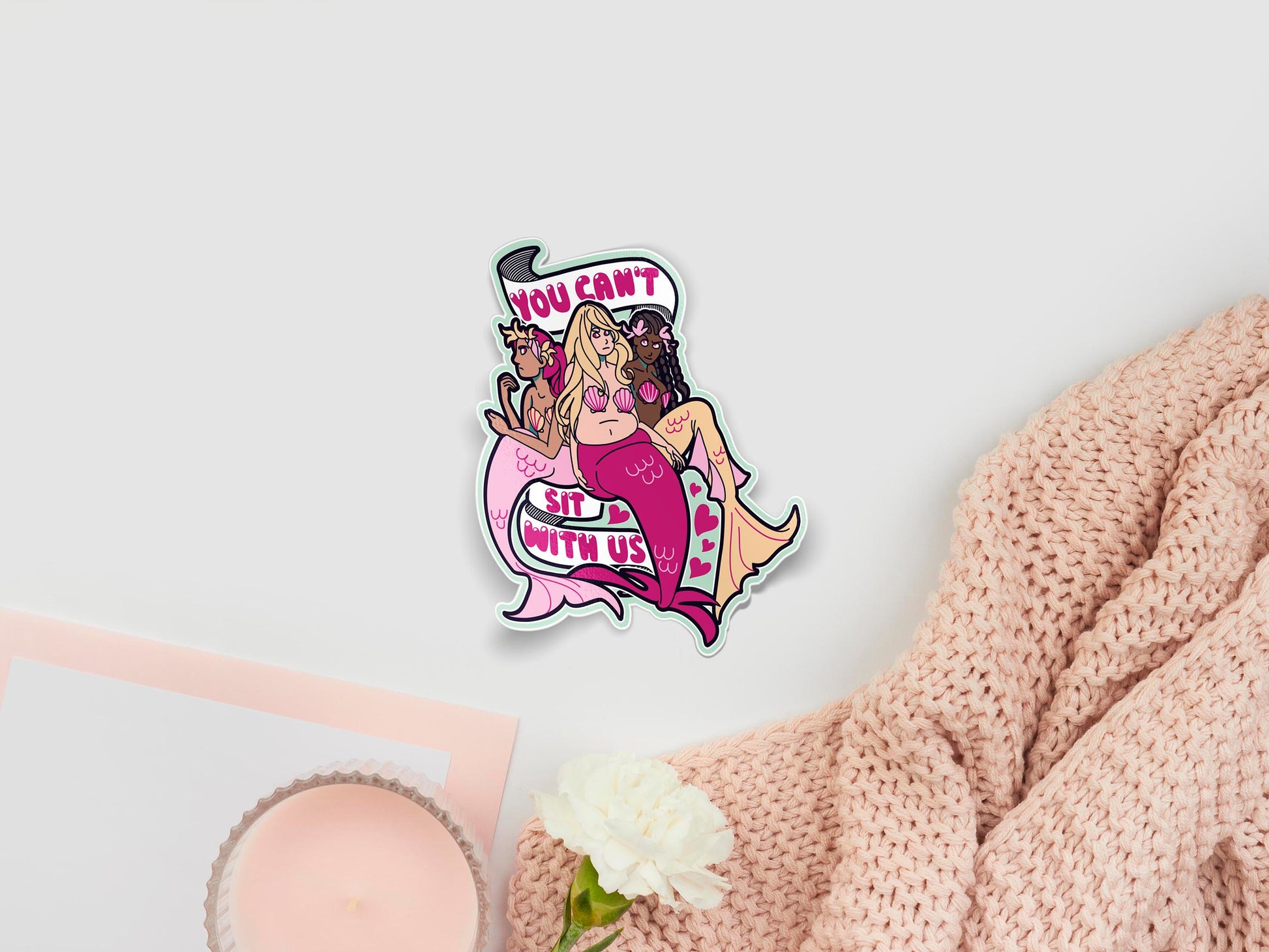 Large sticker sheet of digital illustration cartoons of three sitting mermaids with different skin tones and tail colours, surrounded by hearts and a ribbon with a the quote You Can't Sit With Us