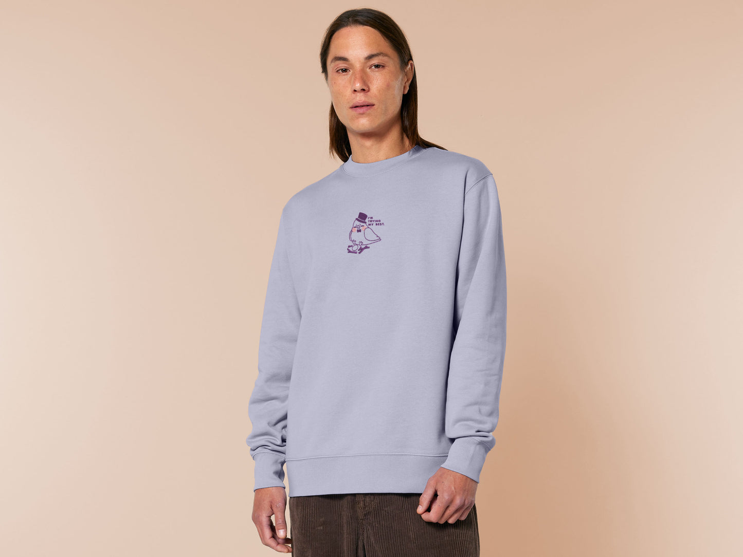 An embroidered purple sweater design of a cute pigeon wearing a top hat building a pathetic nest and the text I'm trying my best.