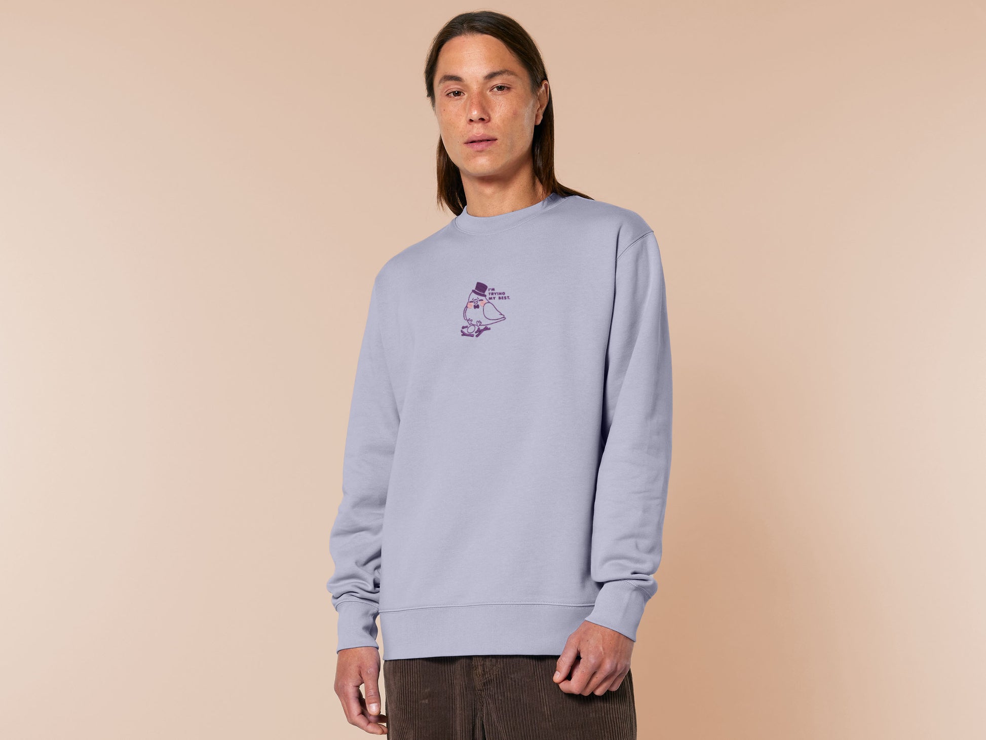 An embroidered purple sweater design of a cute pigeon wearing a top hat building a pathetic nest and the text I'm trying my best.