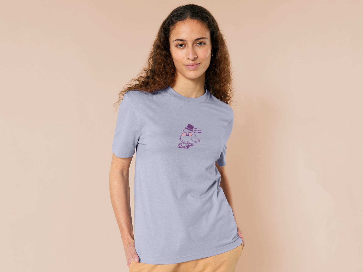 An embroidered purple t-shirt design of a cute pigeon wearing a top hat building a pathetic nest and the text I'm trying my best.