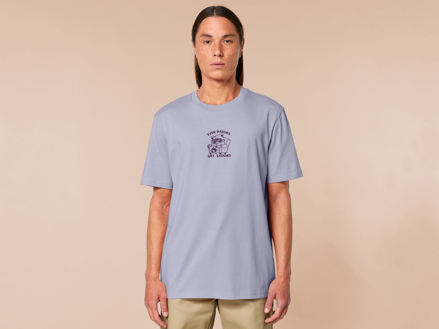 Purple embroidered cute t-shirt design of a chibi raccoon crying wrapped in a blanket sat on a sofa with the quote Even Baddies Get Saddies