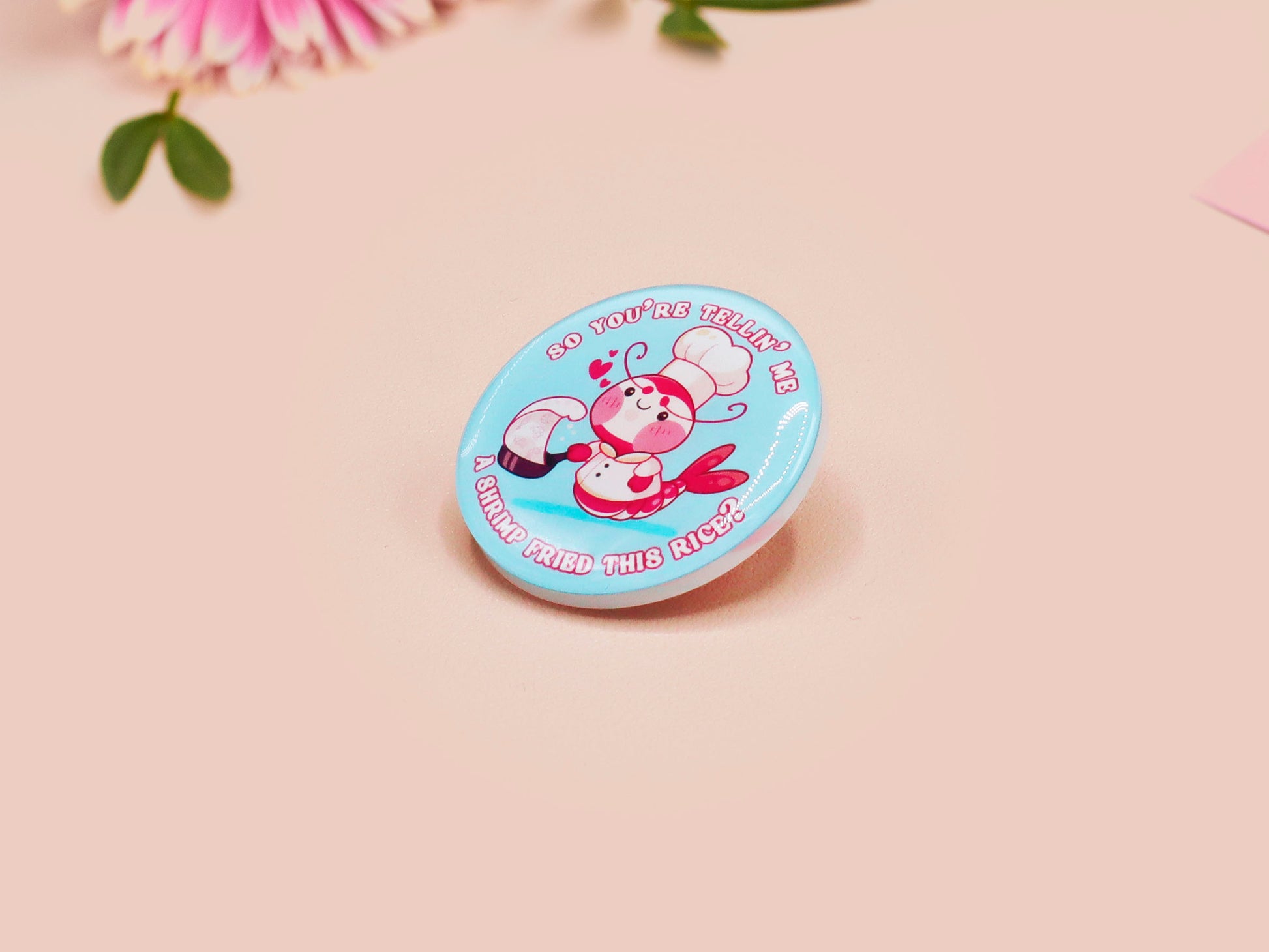 A clear acrylic pin badge with a pink heart-shaped pin back clasp. The design of the pin badge is of a cute chibi shrimp in a chefs outfit cooking with the quote So you're telling me a shrimp fried this rice?