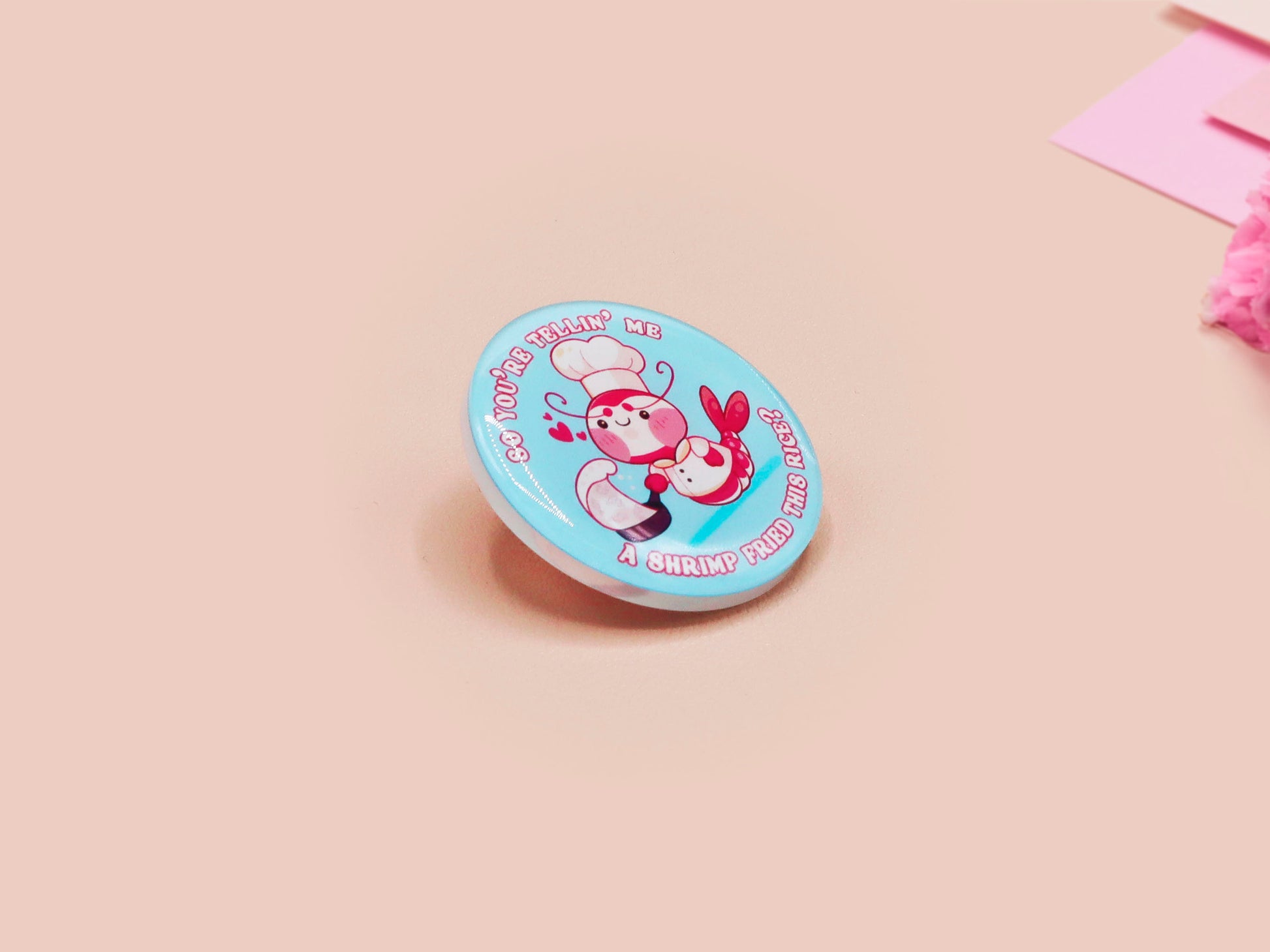 A clear acrylic pin badge with a pink heart-shaped pin back clasp. The design of the pin badge is of a cute chibi shrimp in a chefs outfit cooking with the quote So you're telling me a shrimp fried this rice?