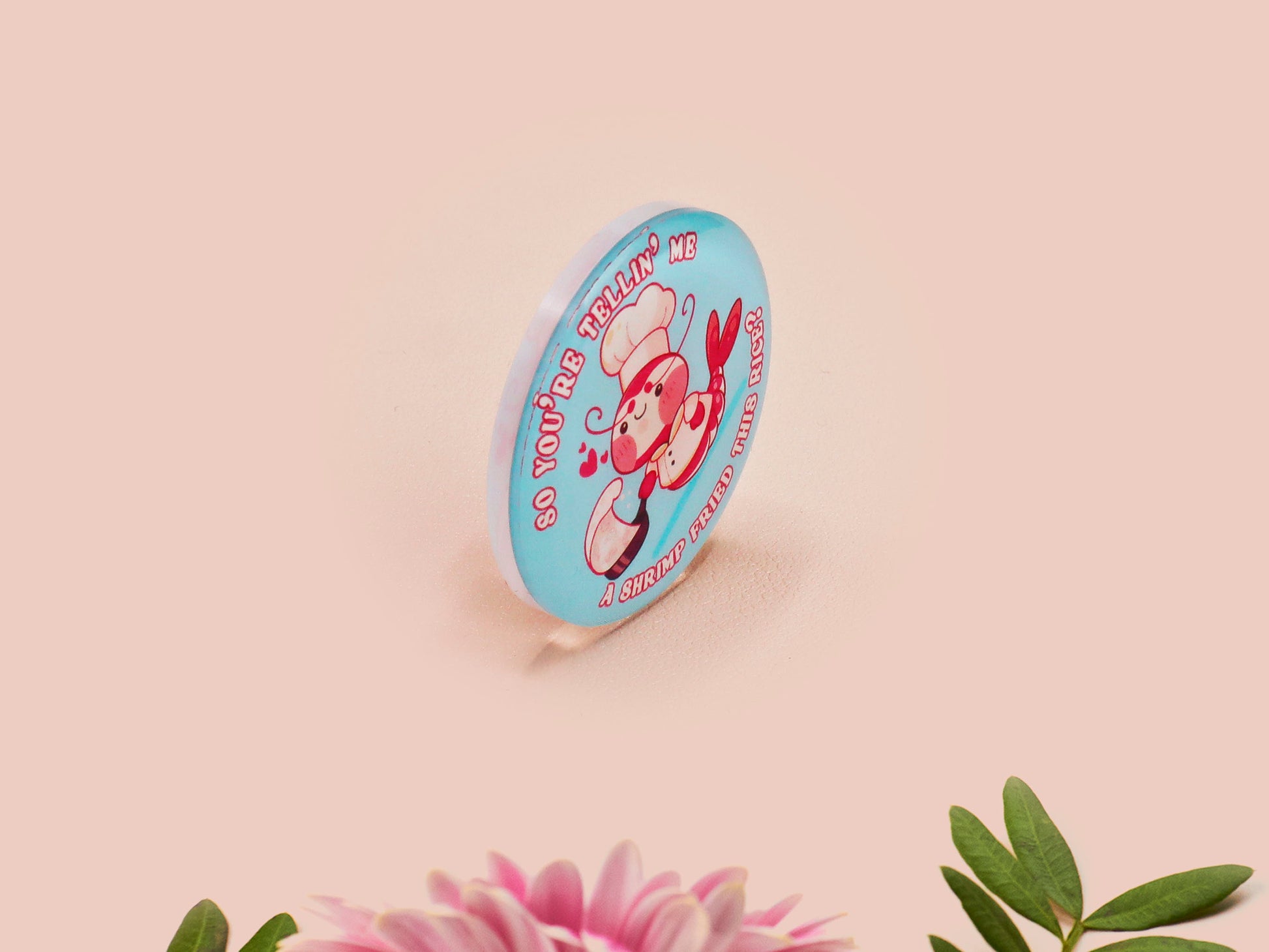 A clear acrylic pin badge with a pink heart-shaped pin back clasp. The design of the pin badge is of a cute chibi shrimp in a chefs outfit cooking with the quote So you're telling me a shrimp fried this rice?