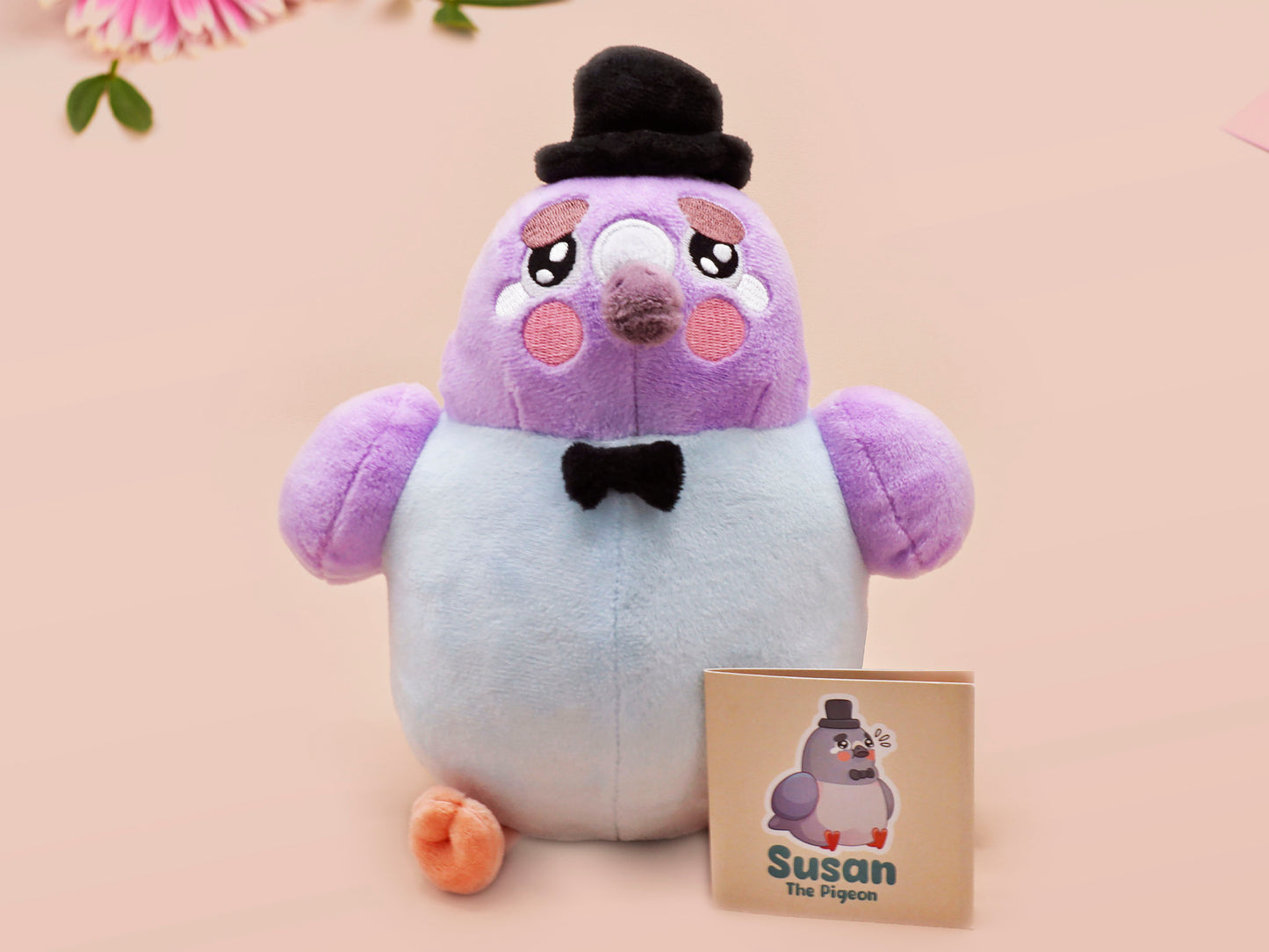 An adorable purple and blue pigeon bird plushie toy with cute embroidered eyes and facial expression, wearing a black top hat and bow tie, with a hang tag describing the pigeon as Susan.