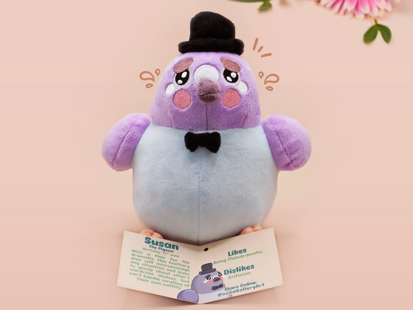 An adorable purple and blue pigeon bird plushie toy with cute embroidered eyes and facial expression, wearing a black top hat and bow tie, with a hang tag describing the pigeon as Susan.