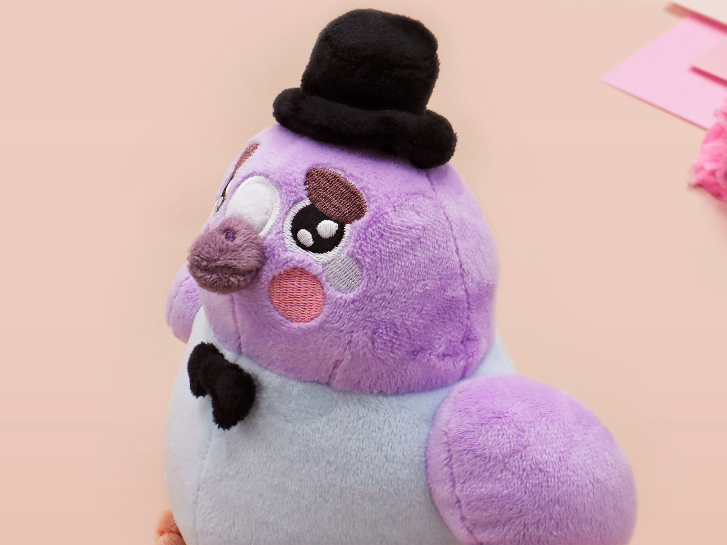 An adorable purple and blue pigeon bird plushie toy with cute embroidered eyes and facial expression, wearing a black top hat and bow tie, with a hang tag describing the pigeon as Susan.