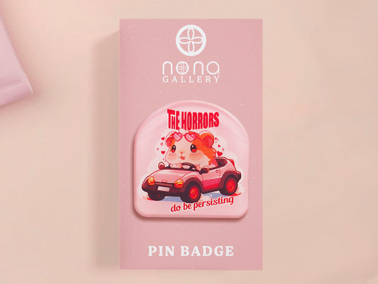 Clear acrylic pin badge with a pink heart-shaped rubber pin back clasp. The design of the pin badge is a cute chibi guinea pig hamster driving a pink car and the quote the horrors do be persisting.