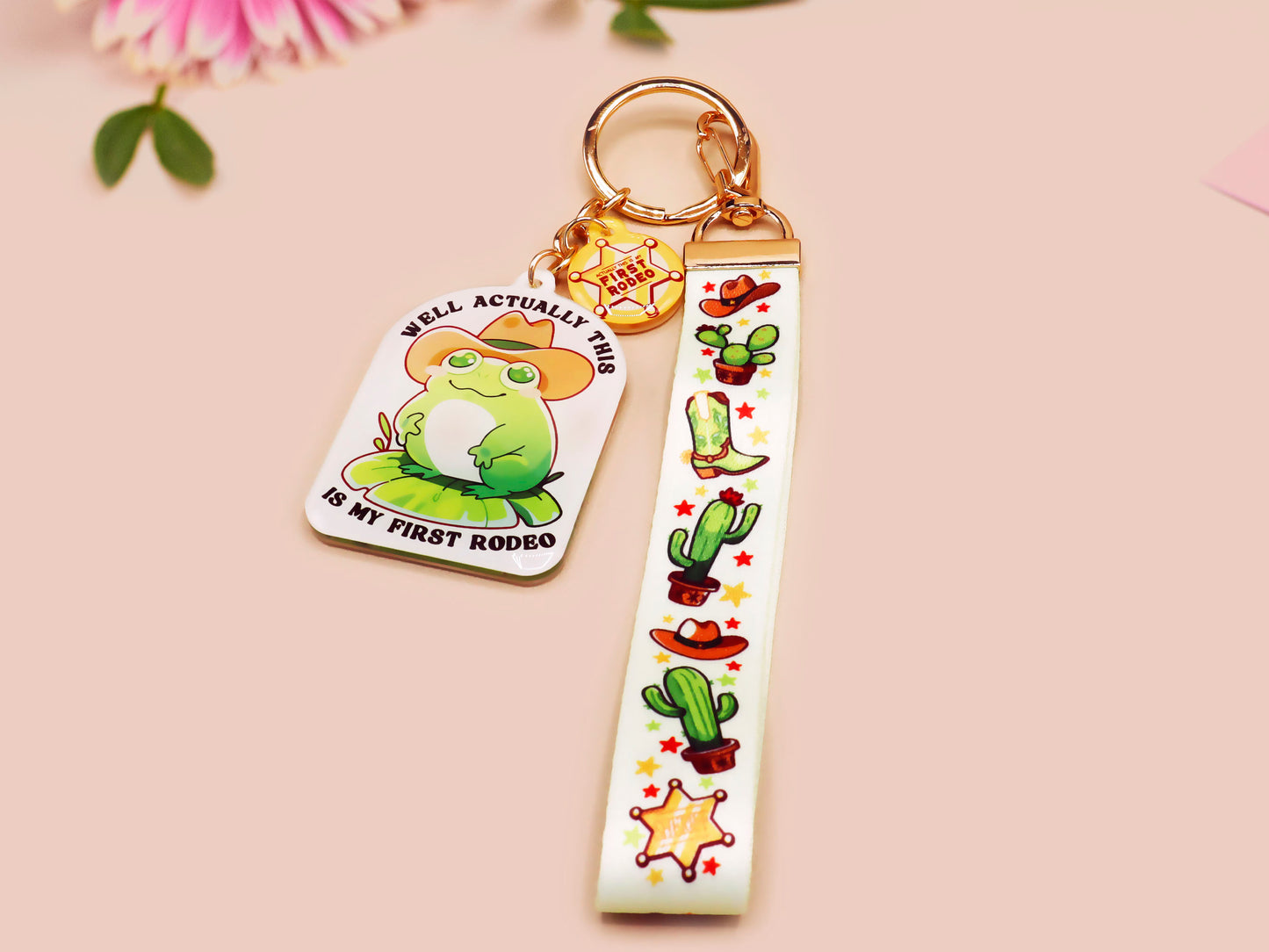 Acrylic epoxy cowboy frog keychain with a large acrylic charm of a cute cowboy frog wearing a stetson hat with the quote well actually this is my first rodeo, a smaller charm of a cute sherrifs badge and a matching cactus and cowboy boots and hats lanyard.