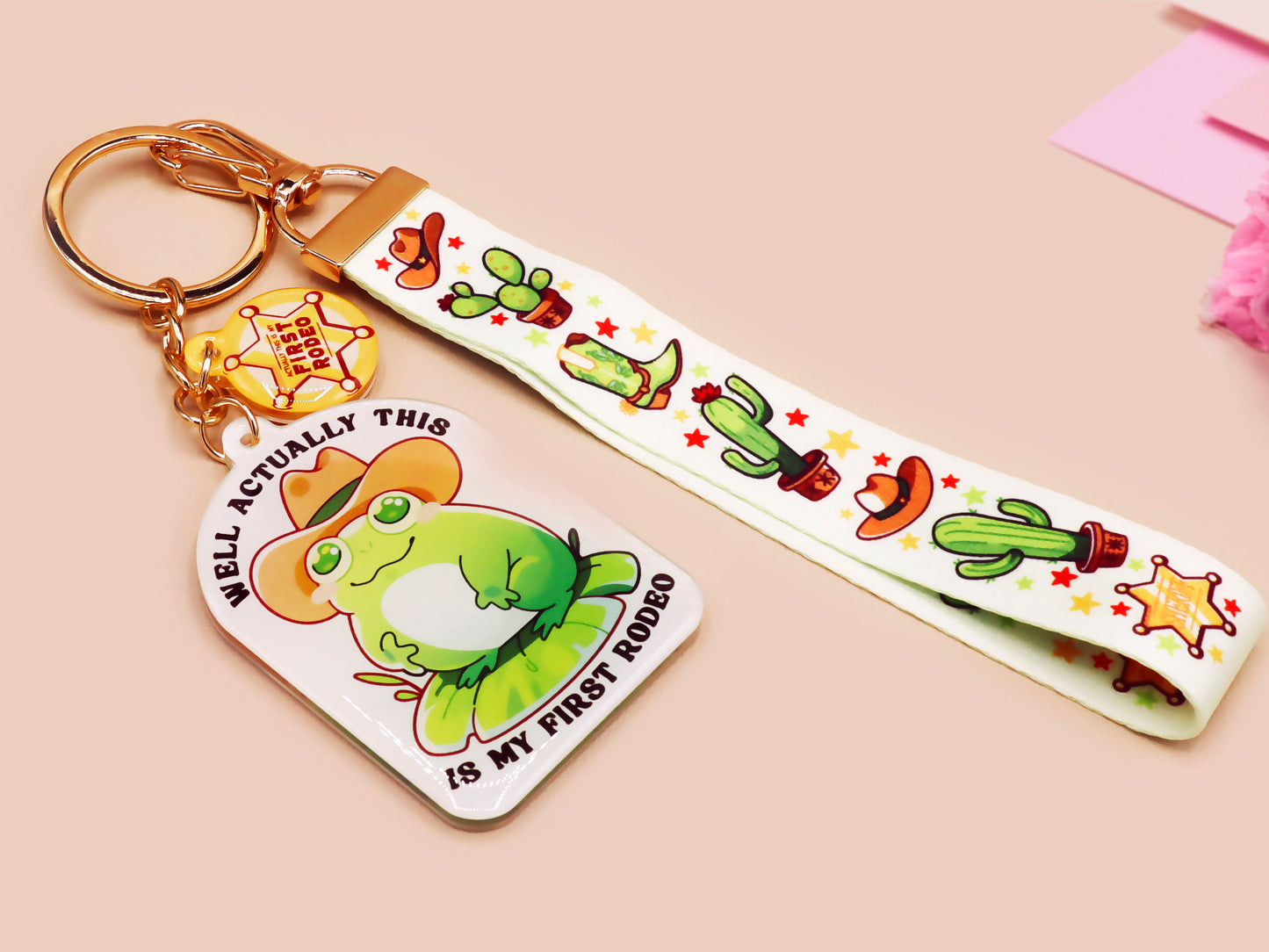 Acrylic epoxy cowboy frog keychain with a large acrylic charm of a cute cowboy frog wearing a stetson hat with the quote well actually this is my first rodeo, a smaller charm of a cute sherrifs badge and a matching cactus and cowboy boots and hats lanyard.