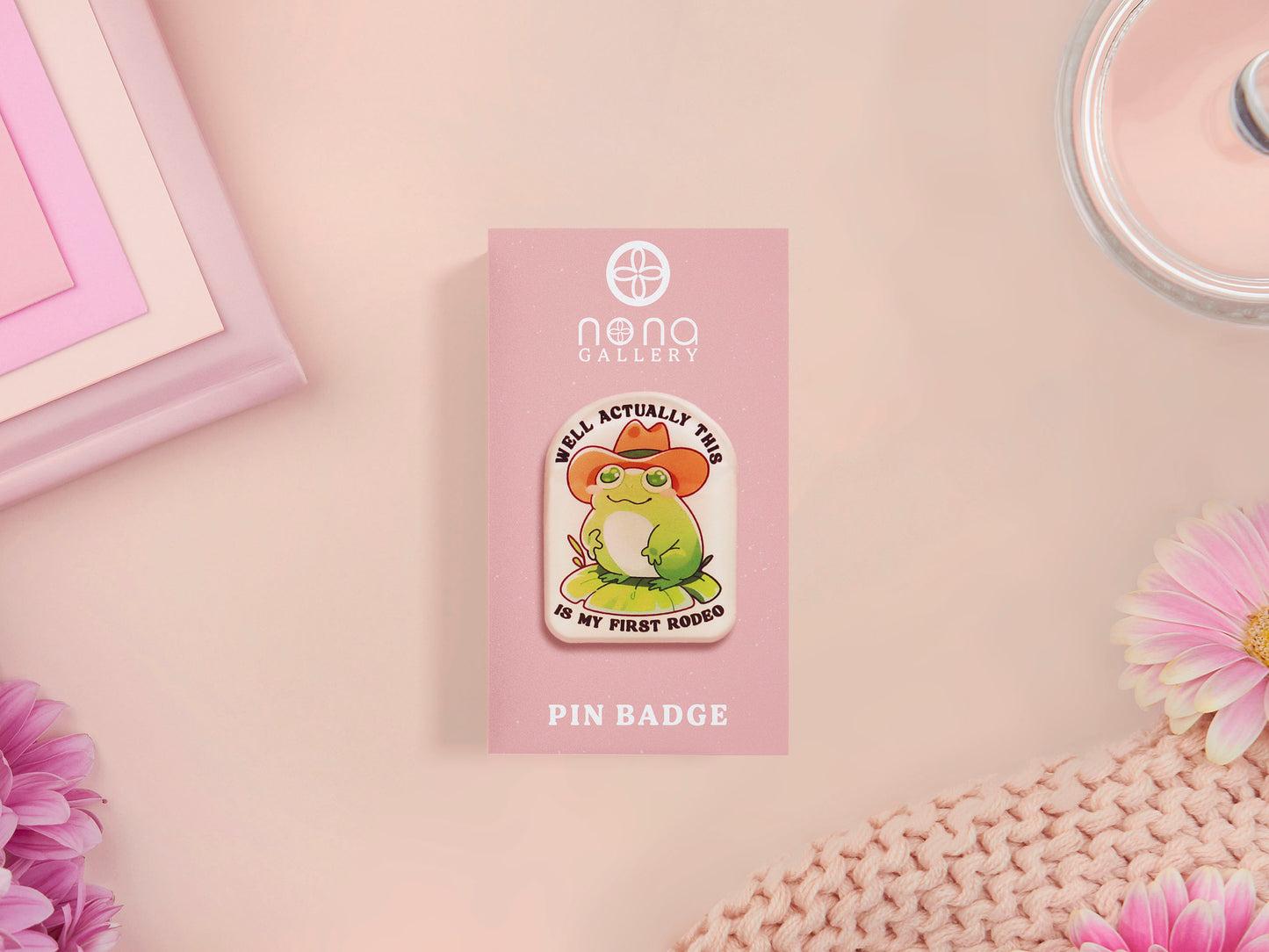 Clear acrylic pin badge with a pink heart-shaped rubber pin back clasp. The design of the pin badge is a cute chibi frog wearing a cowboy hat stetson and the quote well actually this is my first rodeo.
