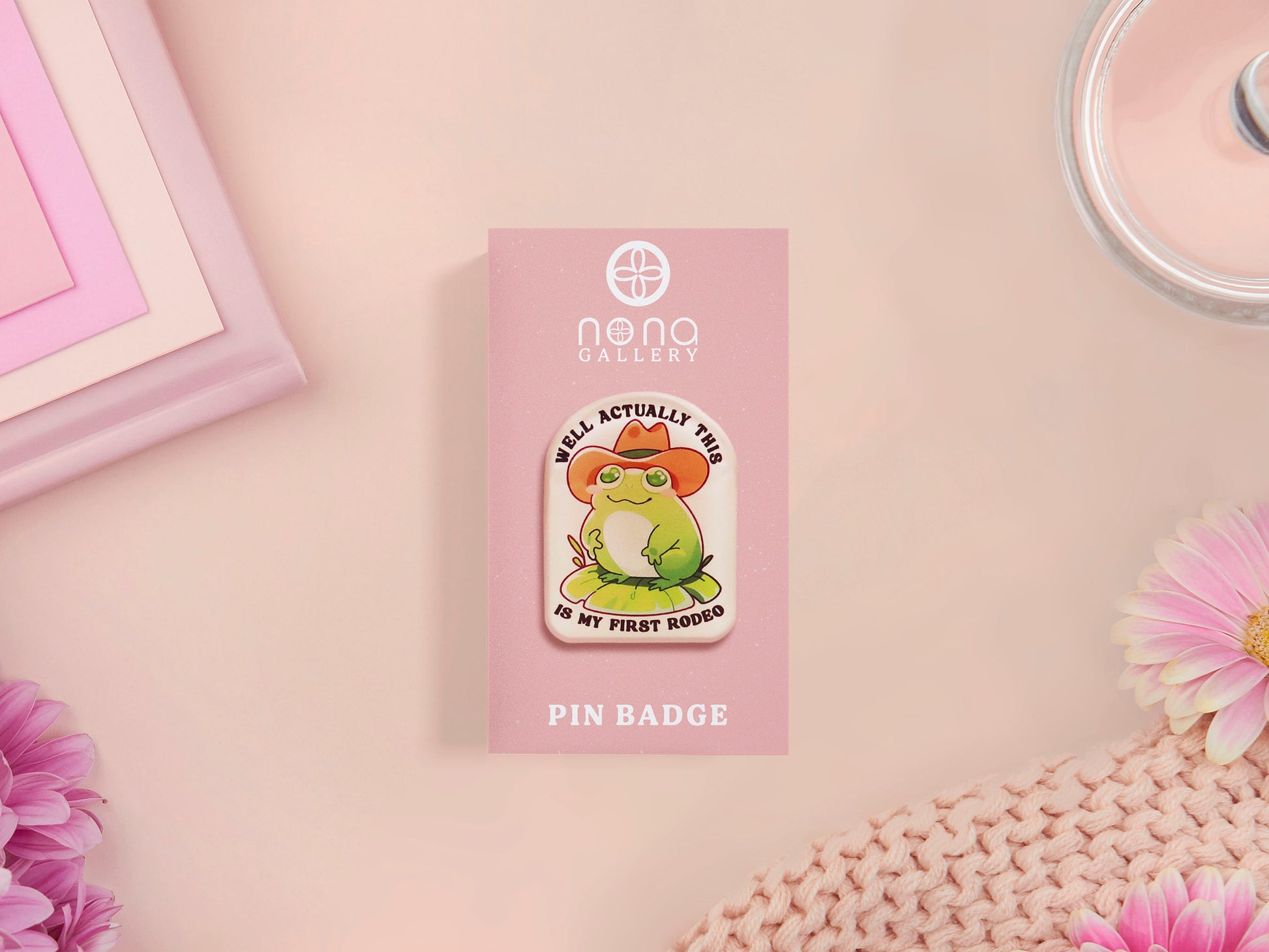 Clear acrylic pin badge with a pink heart-shaped rubber pin back clasp. The design of the pin badge is a cute chibi frog wearing a cowboy hat stetson and the quote well actually this is my first rodeo.