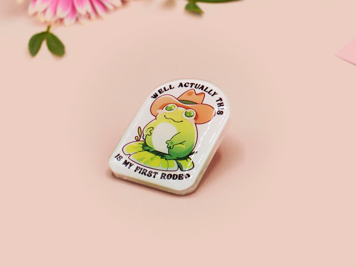 Clear acrylic pin badge with a pink heart-shaped rubber pin back clasp. The design of the pin badge is a cute chibi frog wearing a cowboy hat stetson and the quote well actually this is my first rodeo.