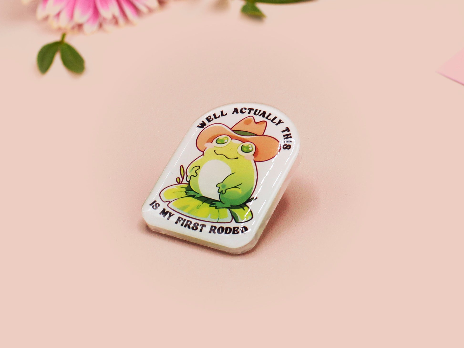 Clear acrylic pin badge with a pink heart-shaped rubber pin back clasp. The design of the pin badge is a cute chibi frog wearing a cowboy hat stetson and the quote well actually this is my first rodeo.