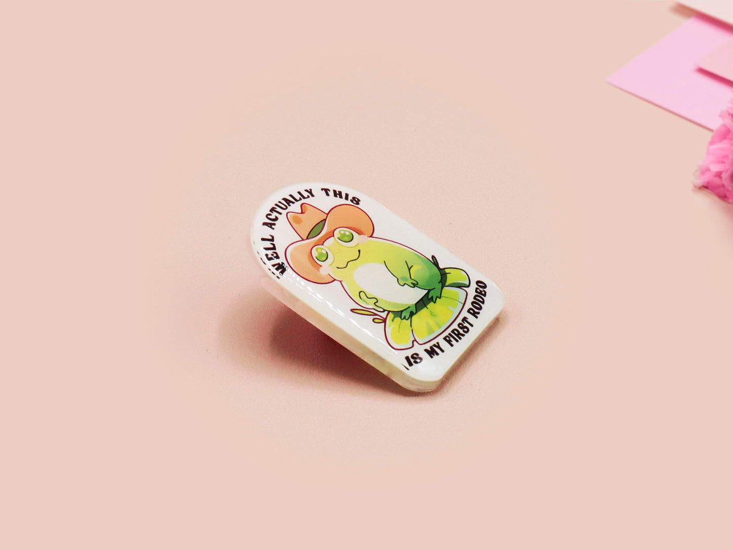 Clear acrylic pin badge with a pink heart-shaped rubber pin back clasp. The design of the pin badge is a cute chibi frog wearing a cowboy hat stetson and the quote well actually this is my first rodeo.