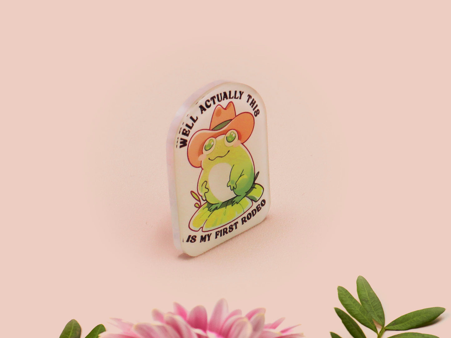 Clear acrylic pin badge with a pink heart-shaped rubber pin back clasp. The design of the pin badge is a cute chibi frog wearing a cowboy hat stetson and the quote well actually this is my first rodeo.