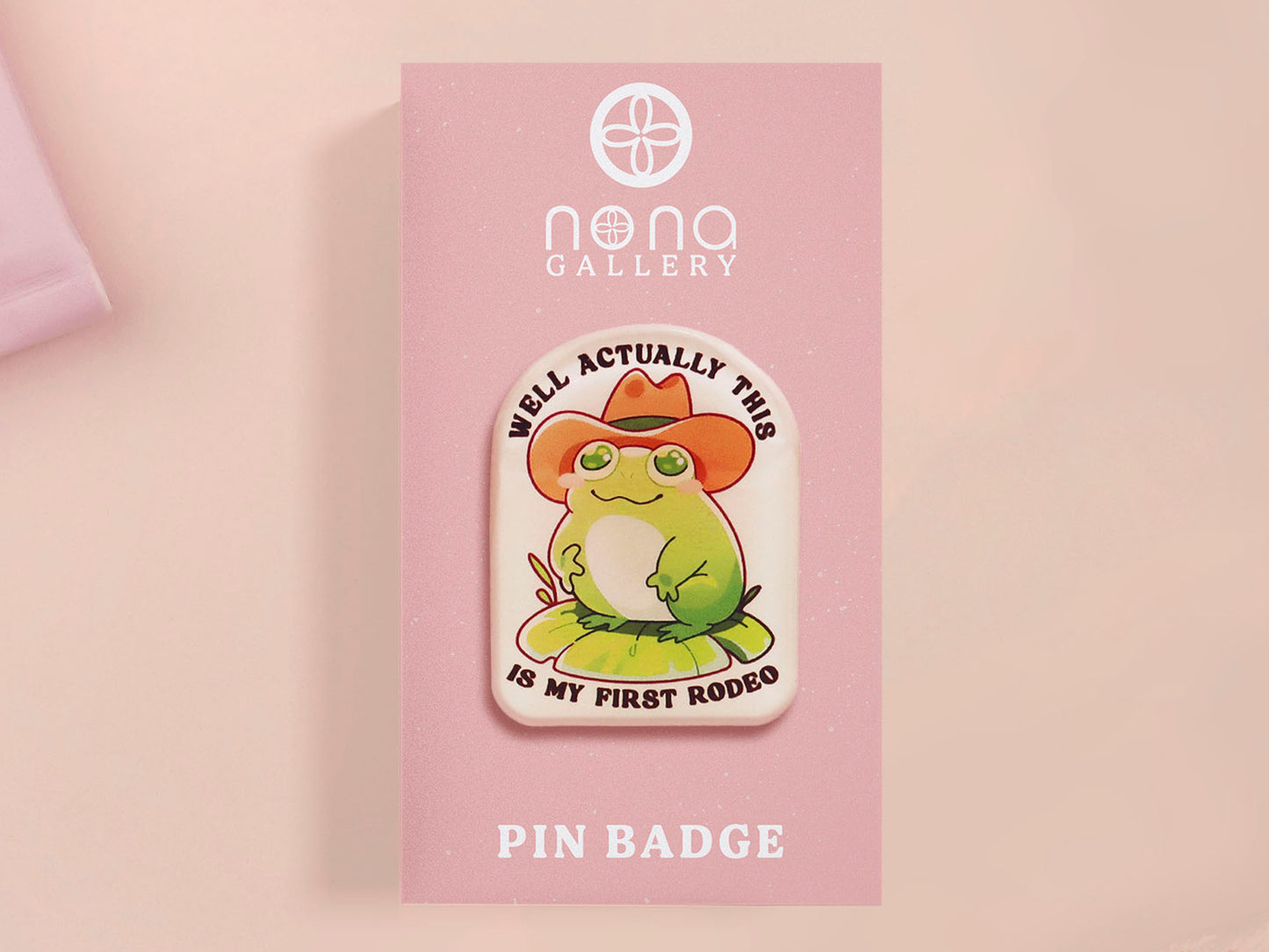 Clear acrylic pin badge with a pink heart-shaped rubber pin back clasp. The design of the pin badge is a cute chibi frog wearing a cowboy hat stetson and the quote well actually this is my first rodeo.