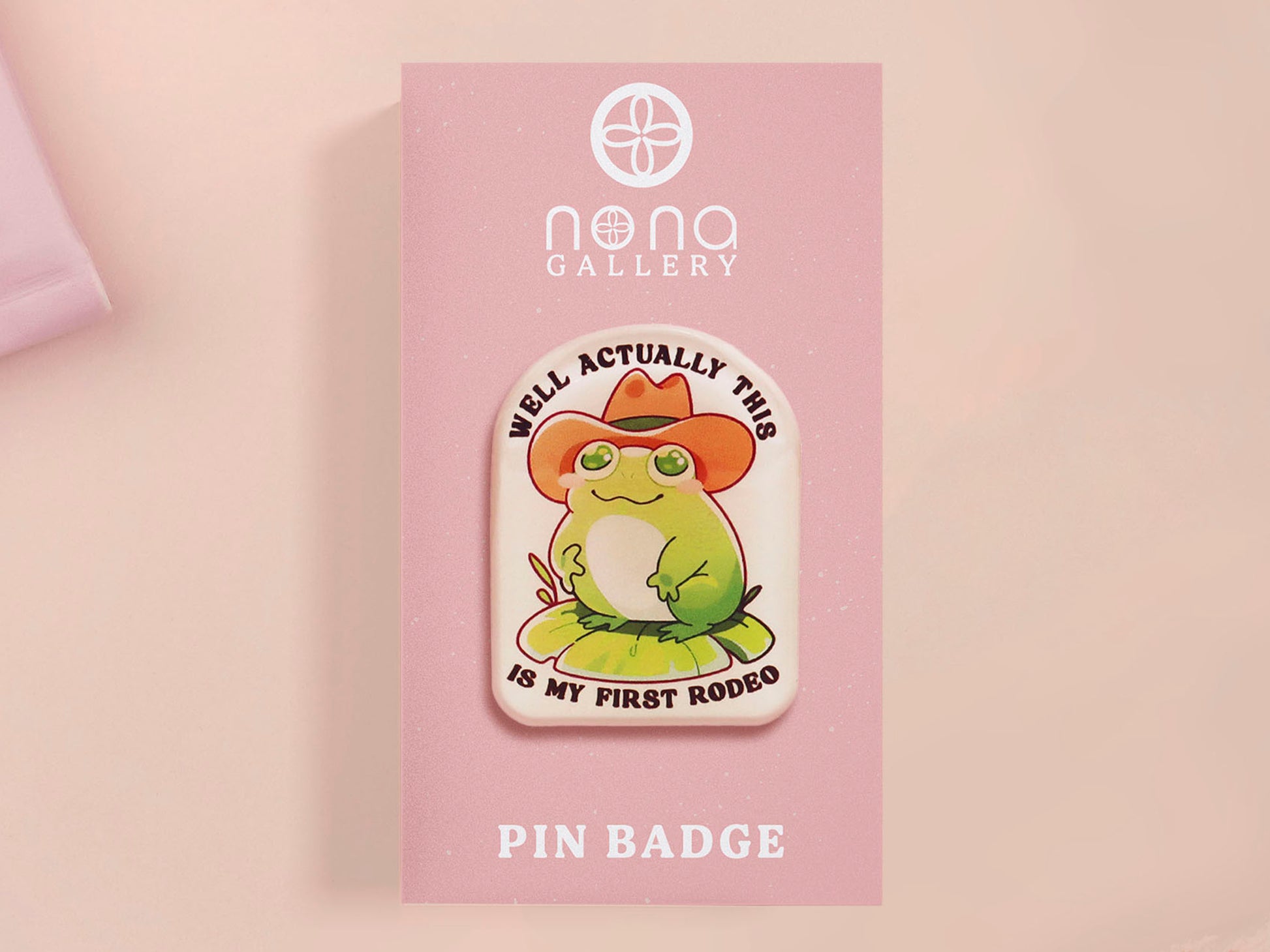 Clear acrylic pin badge with a pink heart-shaped rubber pin back clasp. The design of the pin badge is a cute chibi frog wearing a cowboy hat stetson and the quote well actually this is my first rodeo.