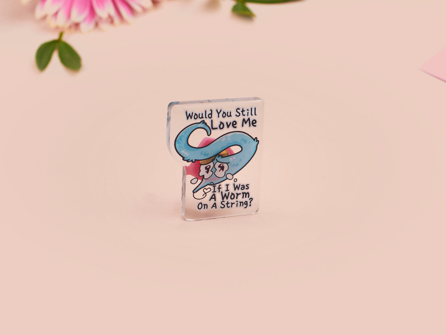 A clear acrylic pin badge with a pink heart-shaped rubber pin back clasp. The pin badge has a design of a cute blue crying worm on a string with the quote would you still love me if I was a worm on a string?