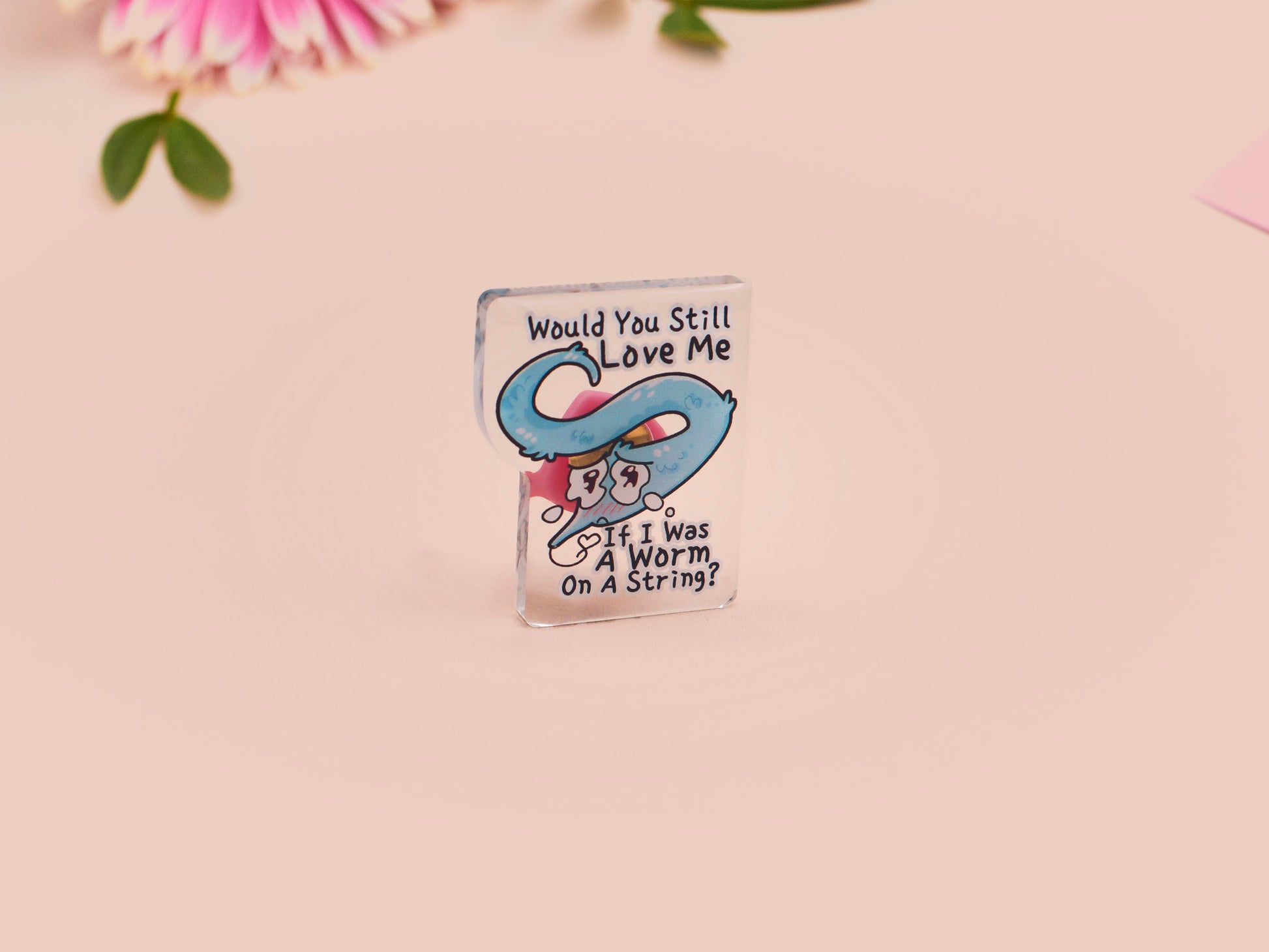 A clear acrylic pin badge with a pink heart-shaped rubber pin back clasp. The pin badge has a design of a cute blue crying worm on a string with the quote would you still love me if I was a worm on a string?