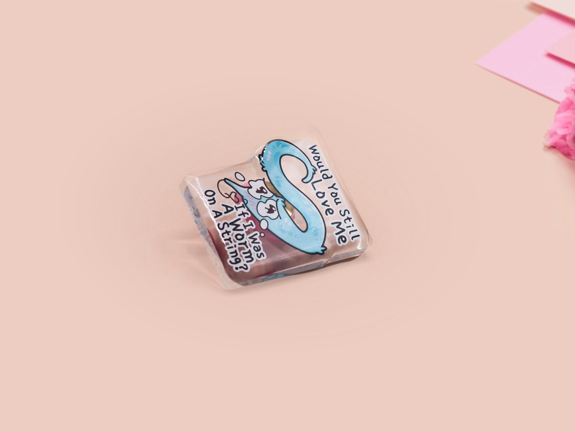 A clear acrylic pin badge with a pink heart-shaped rubber pin back clasp. The pin badge has a design of a cute blue crying worm on a string with the quote would you still love me if I was a worm on a string?
