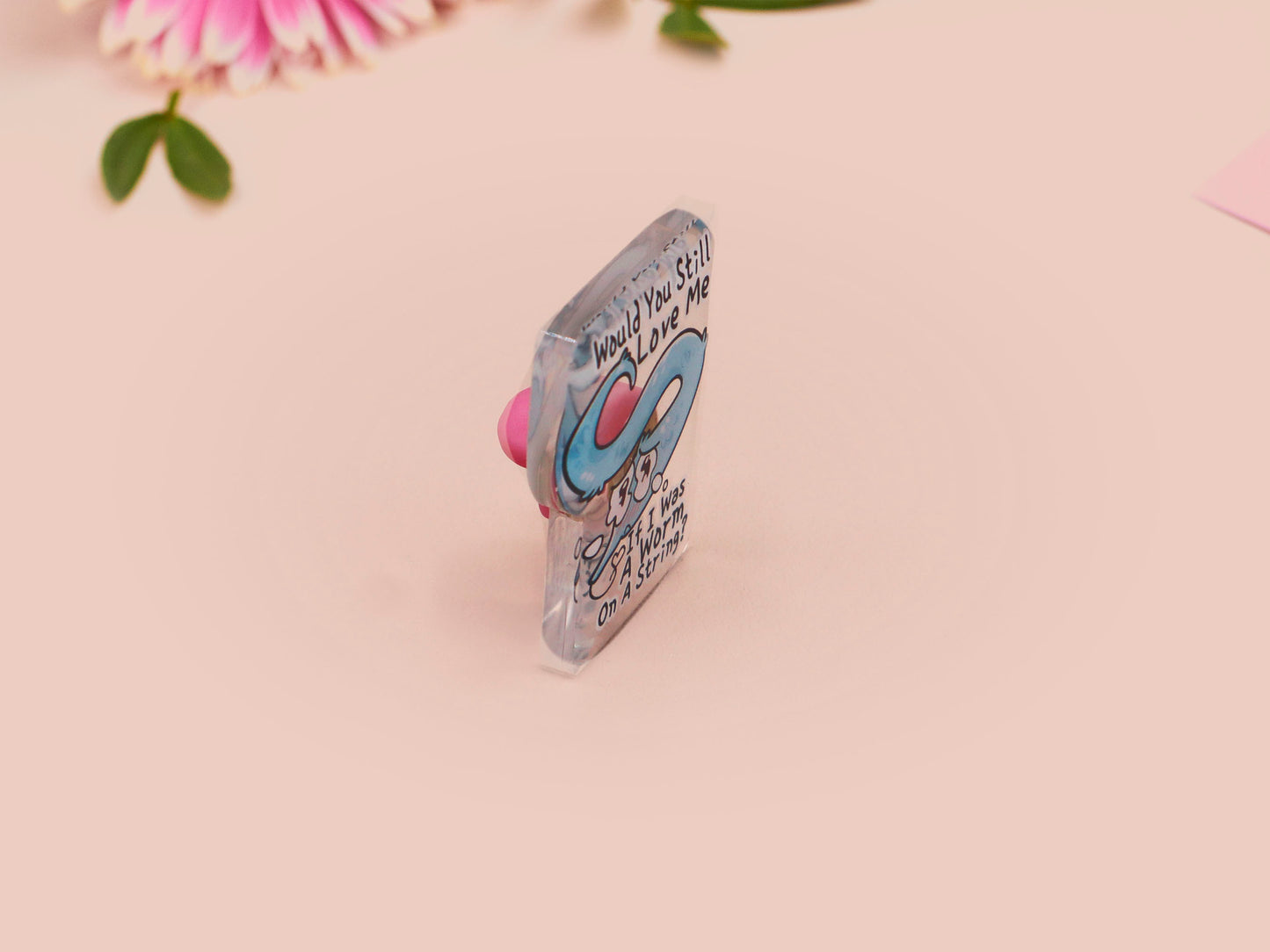 A clear acrylic pin badge with a pink heart-shaped rubber pin back clasp. The pin badge has a design of a cute blue crying worm on a string with the quote would you still love me if I was a worm on a string?