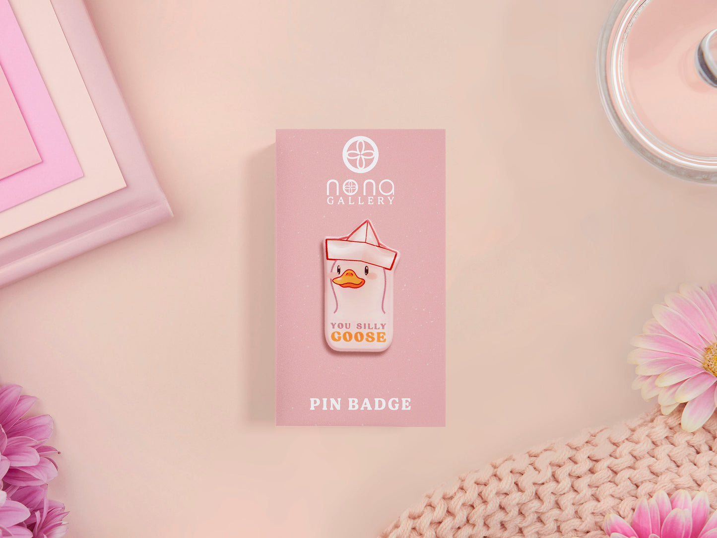 A clear acrylic pin badge with a pink heart-shaped pin back clasp. The design of the pin badge is of a cute chibi goose wearing a paper hat with the quote you silly goose.