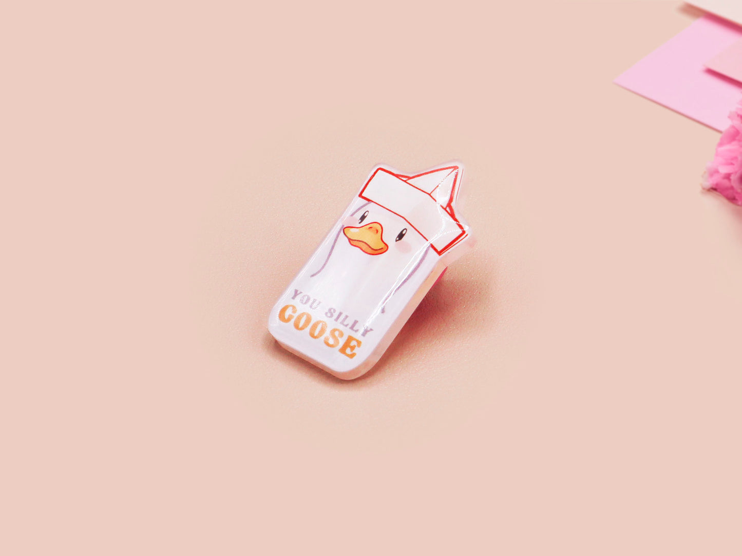 A clear acrylic pin badge with a pink heart-shaped pin back clasp. The design of the pin badge is of a cute chibi goose wearing a paper hat with the quote you silly goose.