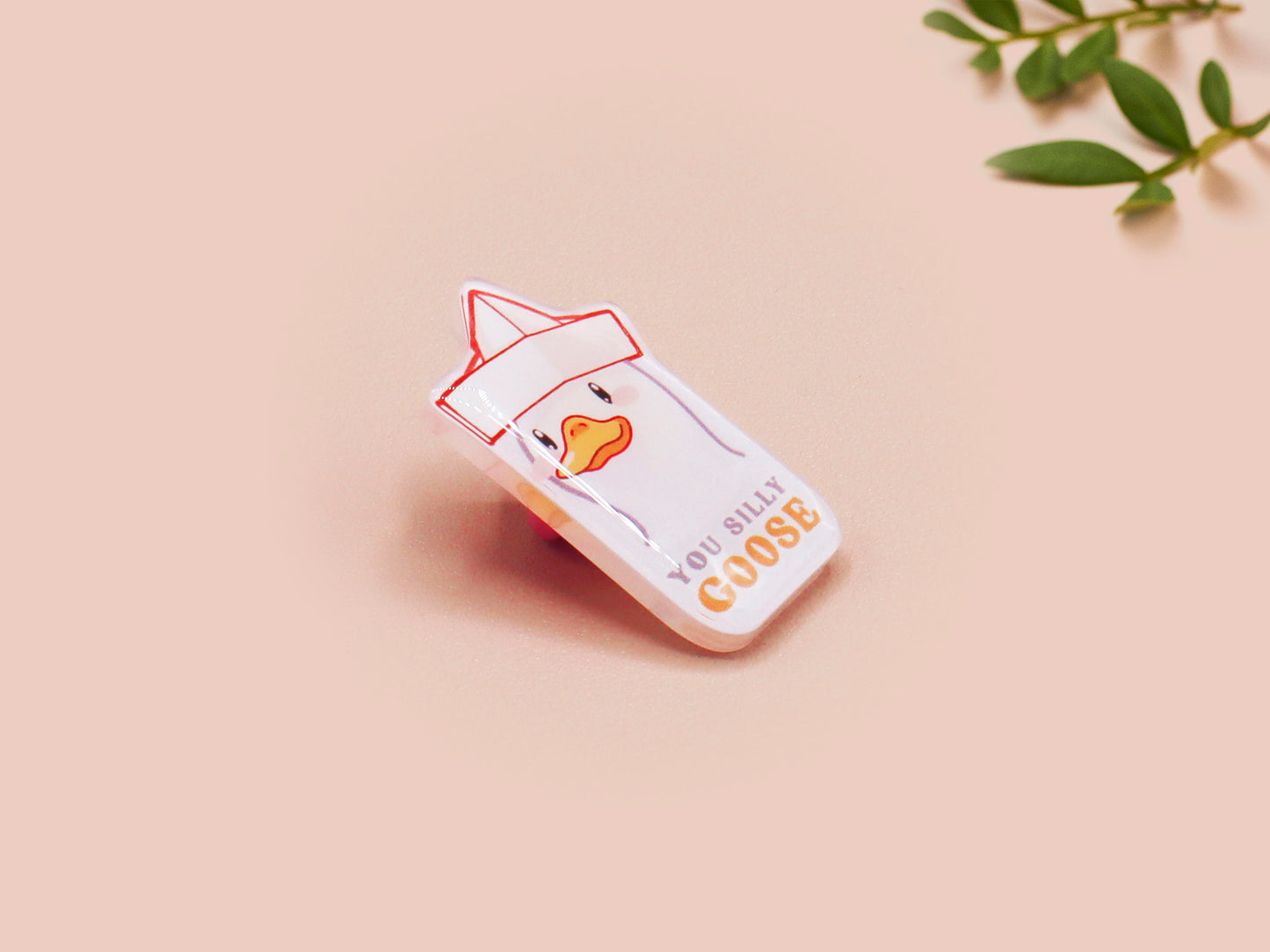 A clear acrylic pin badge with a pink heart-shaped pin back clasp. The design of the pin badge is of a cute chibi goose wearing a paper hat with the quote you silly goose.