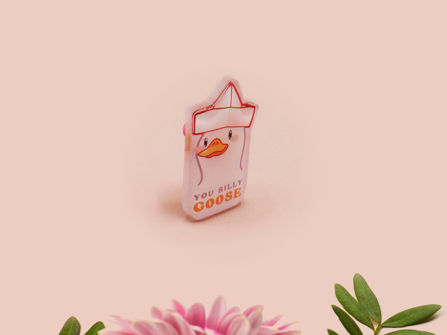 A clear acrylic pin badge with a pink heart-shaped pin back clasp. The design of the pin badge is of a cute chibi goose wearing a paper hat with the quote you silly goose.
