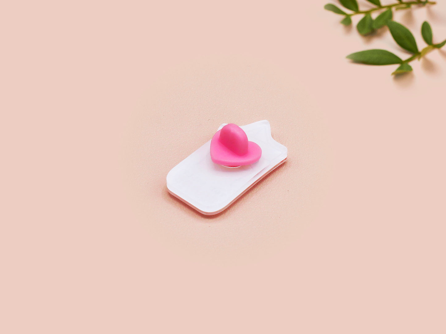 A clear acrylic pin badge with a pink heart-shaped pin back clasp. The design of the pin badge is of a cute chibi goose wearing a paper hat with the quote you silly goose.