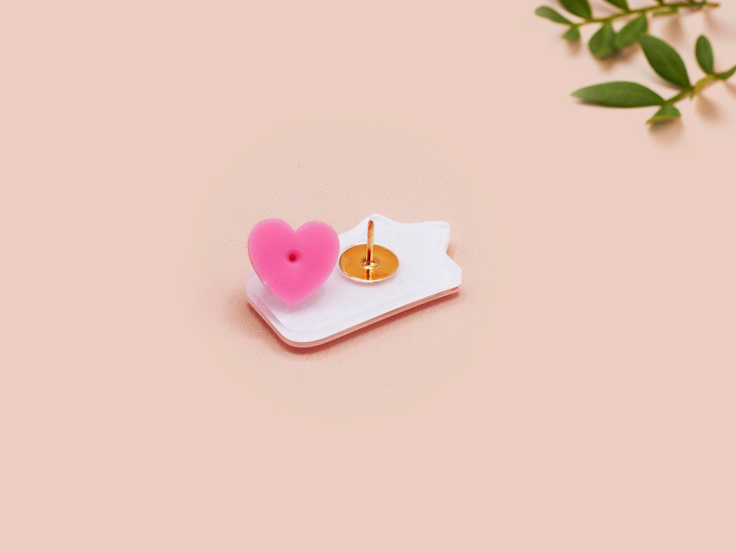 A clear acrylic pin badge with a pink heart-shaped pin back clasp. The design of the pin badge is of a cute chibi goose wearing a paper hat with the quote you silly goose.