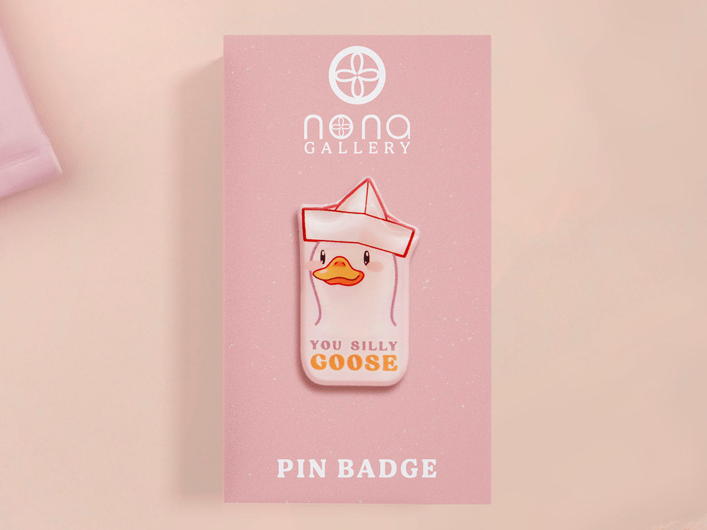 A clear acrylic pin badge with a pink heart-shaped pin back clasp. The design of the pin badge is of a cute chibi goose wearing a paper hat with the quote you silly goose.
