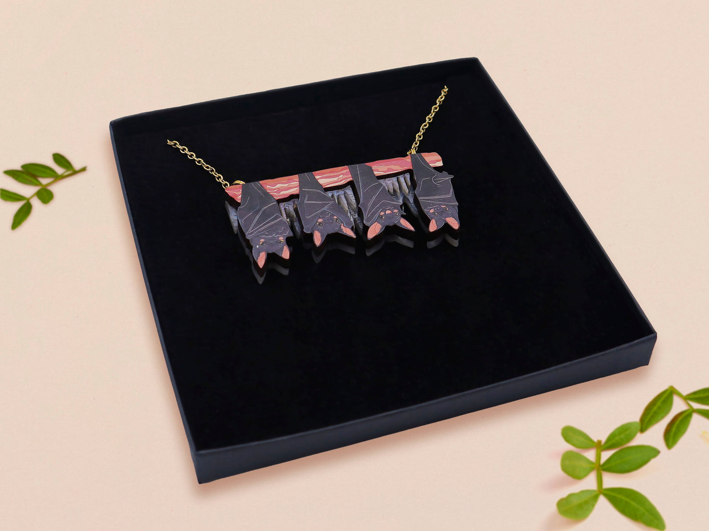 A wooden and glitter acrylic necklace with gold chain in a black box of four cute bats hung upside down on a cave wall of stalactites
