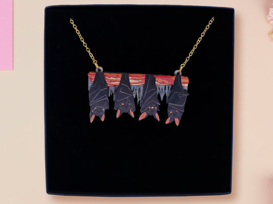 A wooden and glitter acrylic necklace with gold chain in a black box of four cute bats hung upside down on a cave wall of stalactites