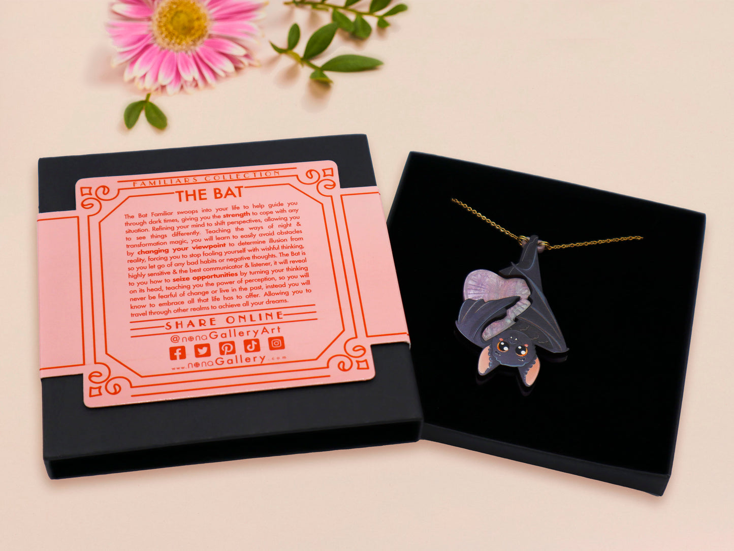 Mixed material handmade necklace of chibi cartoon bat hanging upside down holding a pearlescent Ouija planchette, with a gold chain and black gift box with a pink familiars collection gift sleeve.