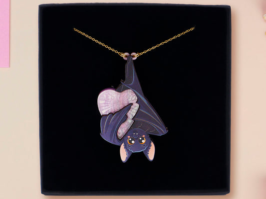 Mixed material handmade necklace of chibi cartoon bat hanging upside down holding a pearlescent Ouija planchette, with a gold chain and black gift box.
