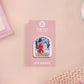 Clear acrylic pin badge with pink heart-shaped rubber pin back clasp. The design of the pin badge is a cute cartoon chibi fresno nightcrawlers, yeti bigfoot and alien UFO in a forest with the quote Be The Cryptid You Want To See In The Woods.