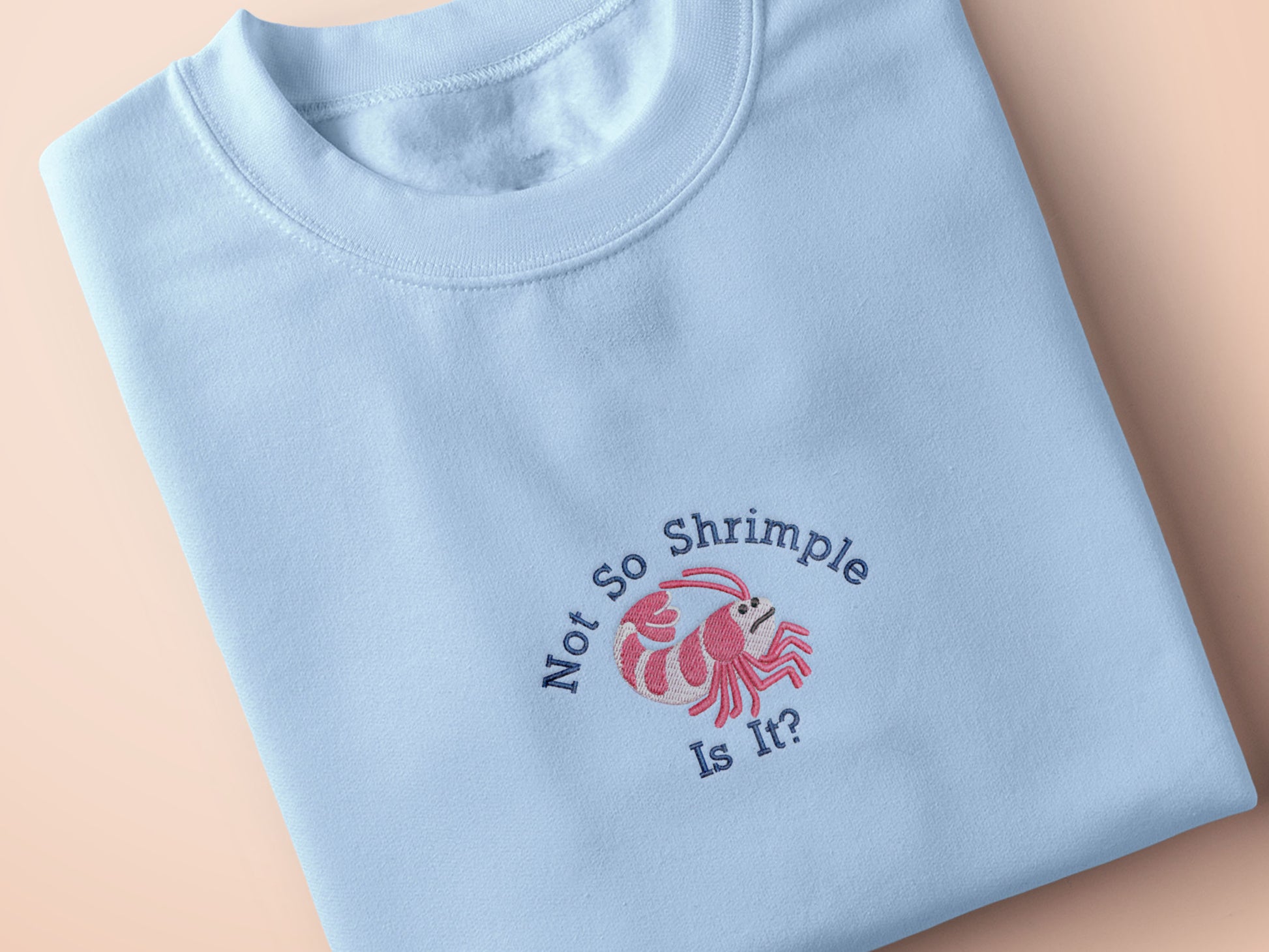 A blue short sleeve tshirt, with an embroidered chibi shrimp looking concerned and the quote Not so shrimple is it?