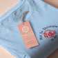 A blue short sleeve tshirt, with an embroidered chibi shrimp looking concerned and the quote Not so shrimple is it?