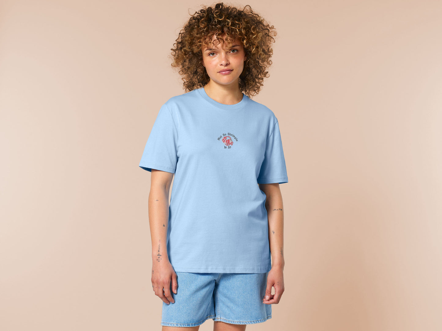 A blue short sleeve tshirt, with an embroidered chibi shrimp looking concerned and the quote Not so shrimple is it?