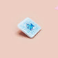 A clear acrylic pin badge with a pink heart-shaped pin back clasp. The design of the pin badge is of a cute chibi blue fish with the quote buy a man eat fish he day teach fish man to a lifetime.